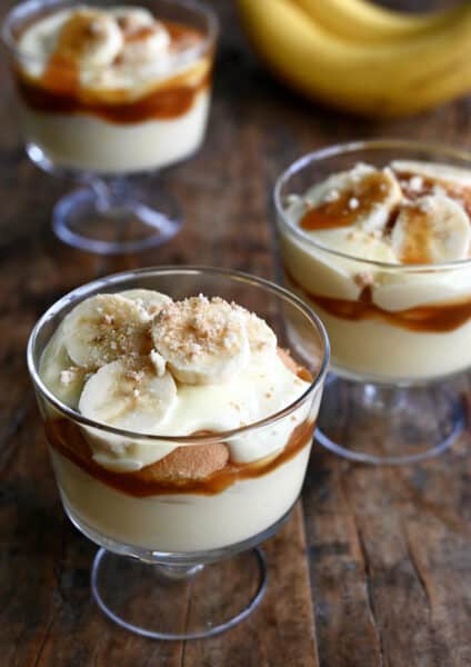 Three parfait glasses each filled with a layer of creamy banana pudding topped with salted caramel sauce, Nilla Wafers, sliced bananas and crushed wafers.
