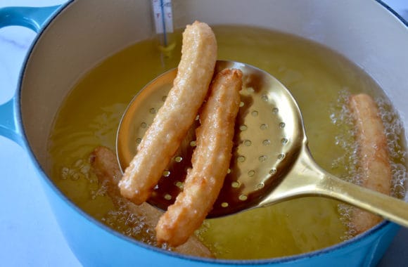 Slotted spoon holding churros over large stockpot containing deep-fry oil