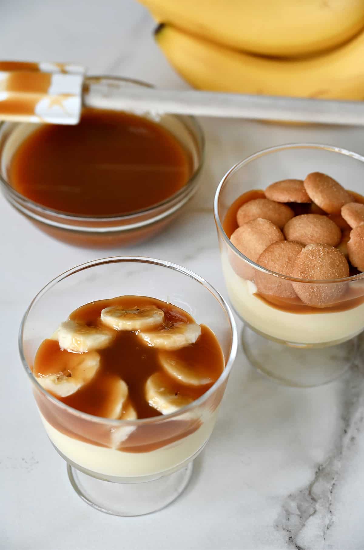 Two parfait glasses containing banana pudding topped with salted caramel sauce, sliced banana and mini vanilla wafers. A glass bowl containing caramel sauce and a spatula is nearby.
