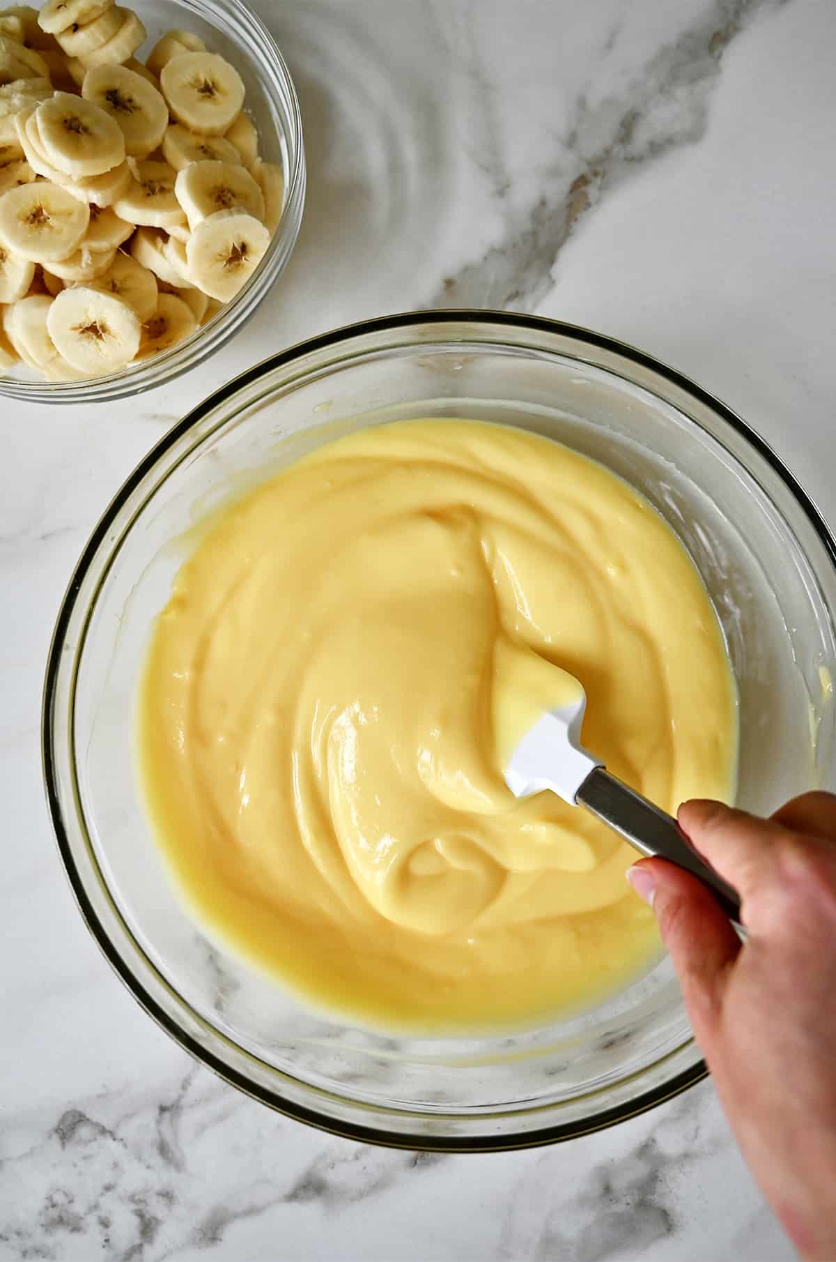 Banana pudding in a glass bowl next to another glass bowl containing fresh banana slices.