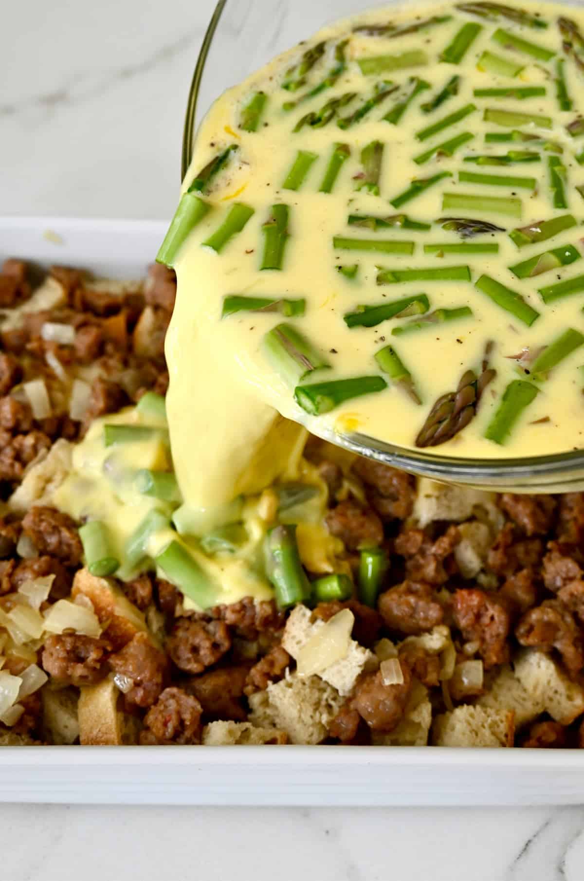 Adding whisked eggs with asparagus and cheese atop bread cubes and cooked sausage crumbles in a 9x13 baking pan.