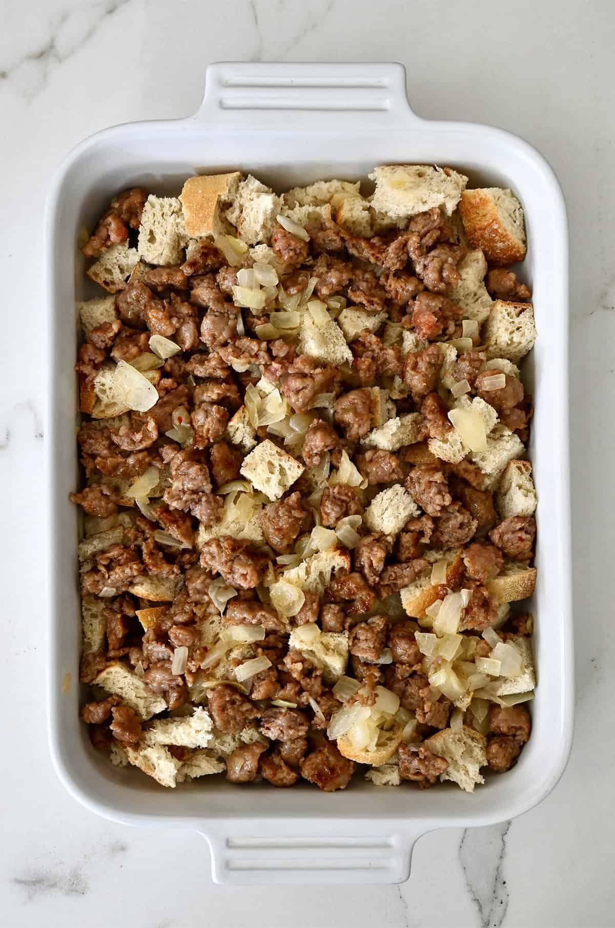 Bread cubes in a 9x13 baking dish with cooked sausage crumbles and diced onions.