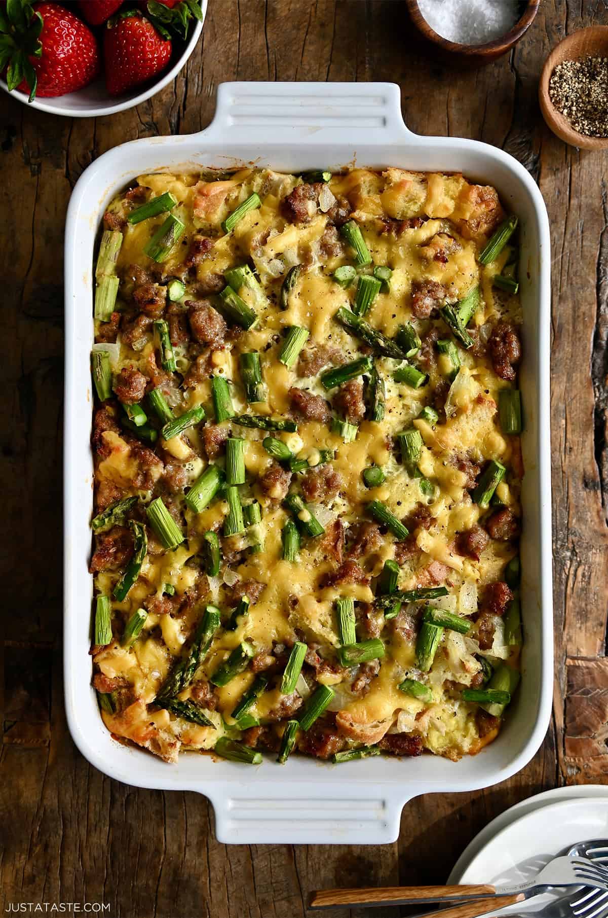A baked overnight breakfast strata in a white baking dish, golden brown on top and filled with layers of breakfast sausage, asparagus and melted cheddar cheese.