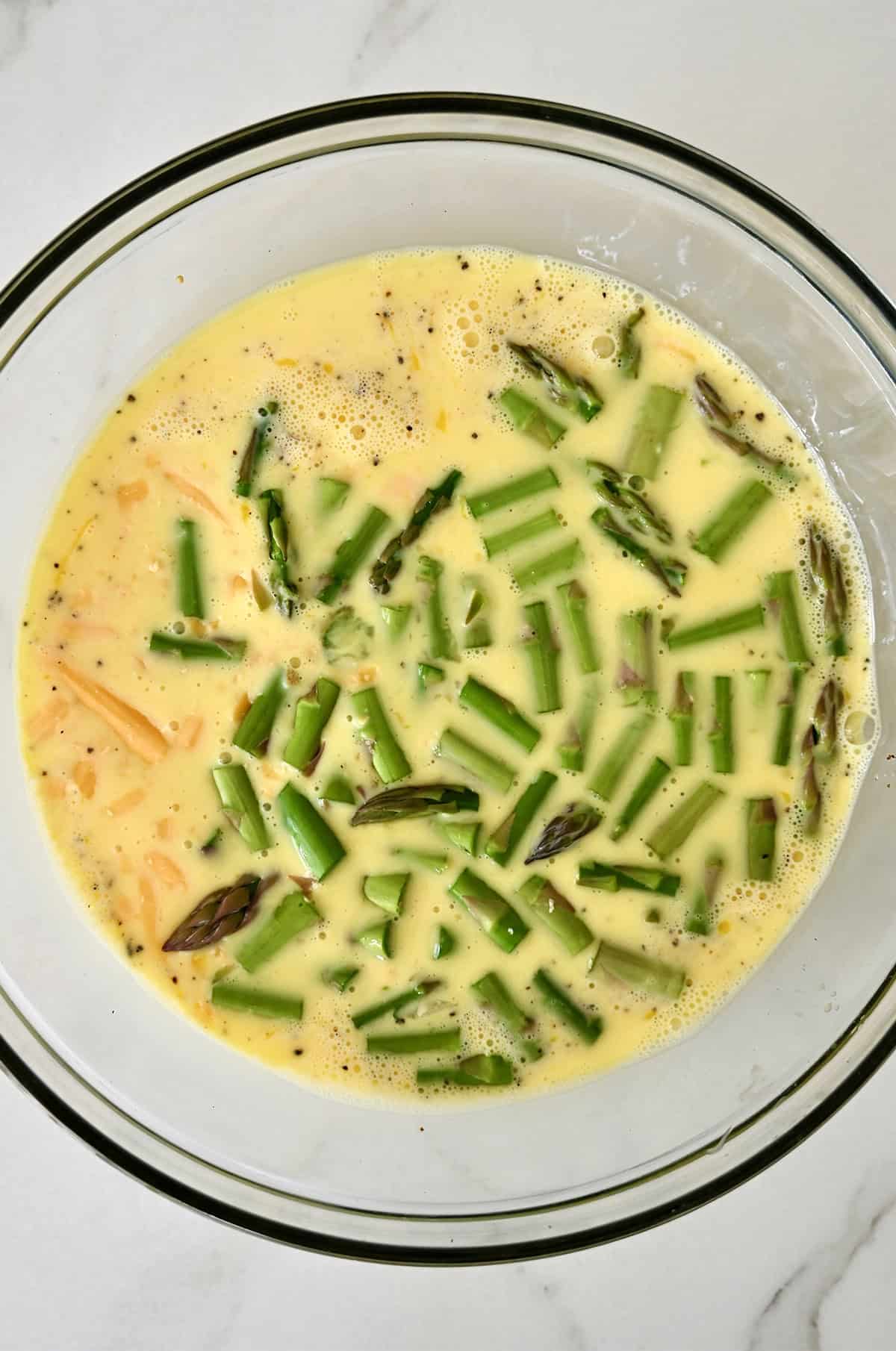 Whisked eggs with milk in a large bowl with shredded cheddar cheese and cut asparagus.