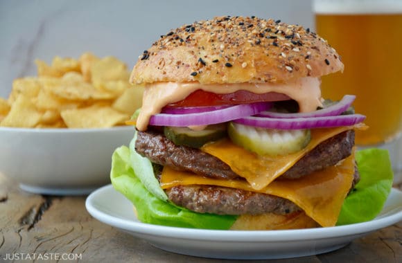 A white plate containing a cheeseburger with chips and a beer in the background