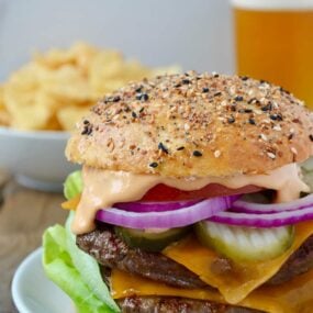 A cheeseburger on a white plate with chips and a beer in the background