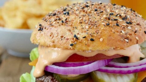 A cheeseburger on a white plate with chips and a beer in the background