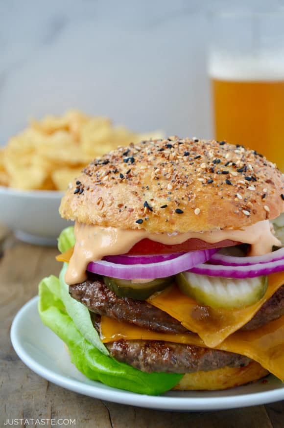 A cheeseburger on a white plate with chips and a beer in the background