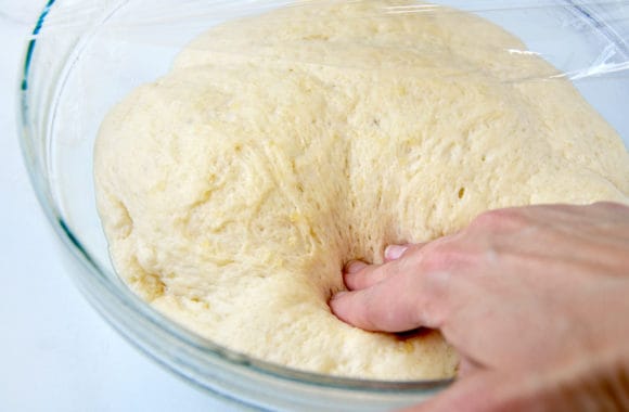 A glass bowl of dough with a hand pushing into it