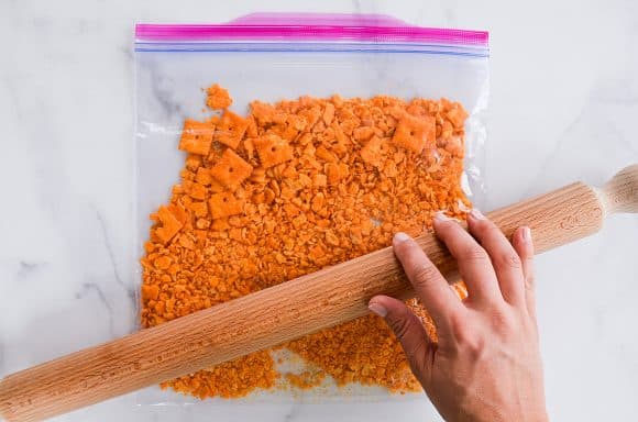 Hand holding rolling pin over plastic bag with crushed cheddar crackers