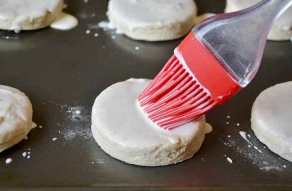 Round shortcake biscuits on nonstick baking sheet being brushed with heavy cream.