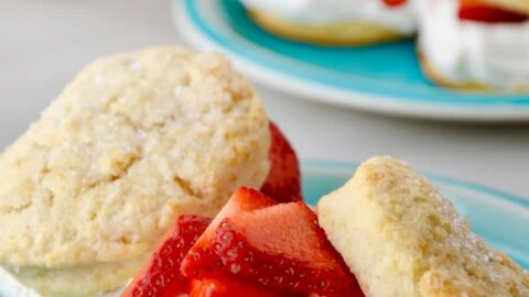 Easy Strawberry Shortcake on blue plate with wooden bowl filled with strawberries and more shortcakes on a blue plate in background.