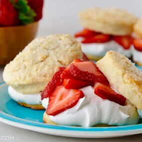 Easy Strawberry Shortcakes with homemade whipped cream on blue plate with small wooden bowl filled with fresh strawberries in background.