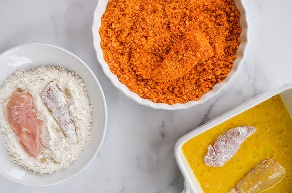 Bowl with chicken fingers and flour next to bowl containing cheddar crackers next to a dish with eggs