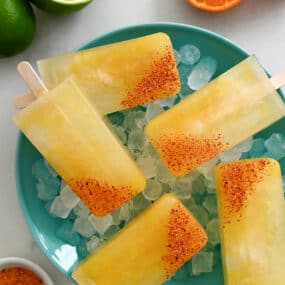 A top-down view of Skinny Margarita Popsicles dipped in tajin seasoning over ice in a bowl next to a halved lime and a halved orange