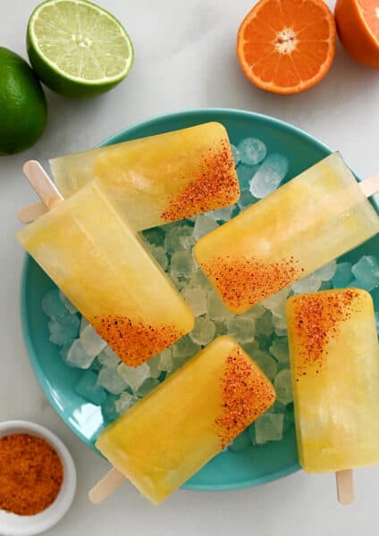 A top-down view of Skinny Margarita Popsicles dipped in tajin seasoning over ice in a bowl next to a halved lime and a halved orange