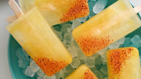 A top-down view of Skinny Margarita Popsicles dipped in tajin seasoning over ice in a bowl next to a halved lime and a halved orange