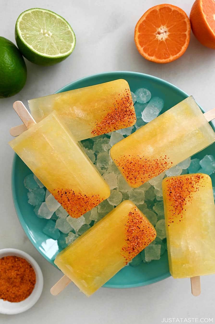 A top-down view of Skinny Margarita Popsicles dipped in tajin seasoning over ice in a bowl next to a halved lime and a halved orange
