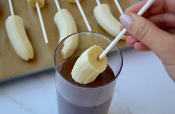 Hand dipping frozen banana in glass containing melted chocolate
