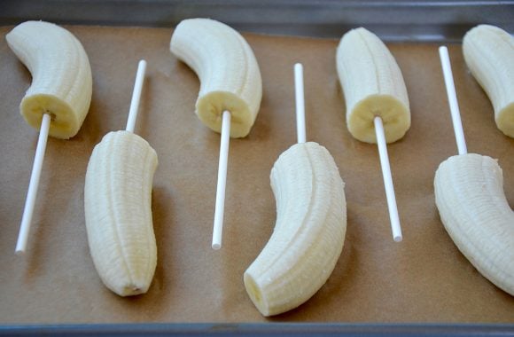 Parchment paper-lined baking sheet with halved bananas with lollipop sticks
