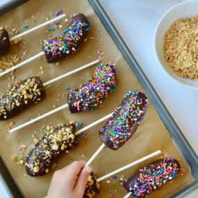 Top down view of child's hand grabbing a frozen banana with chocolate and sprinkles