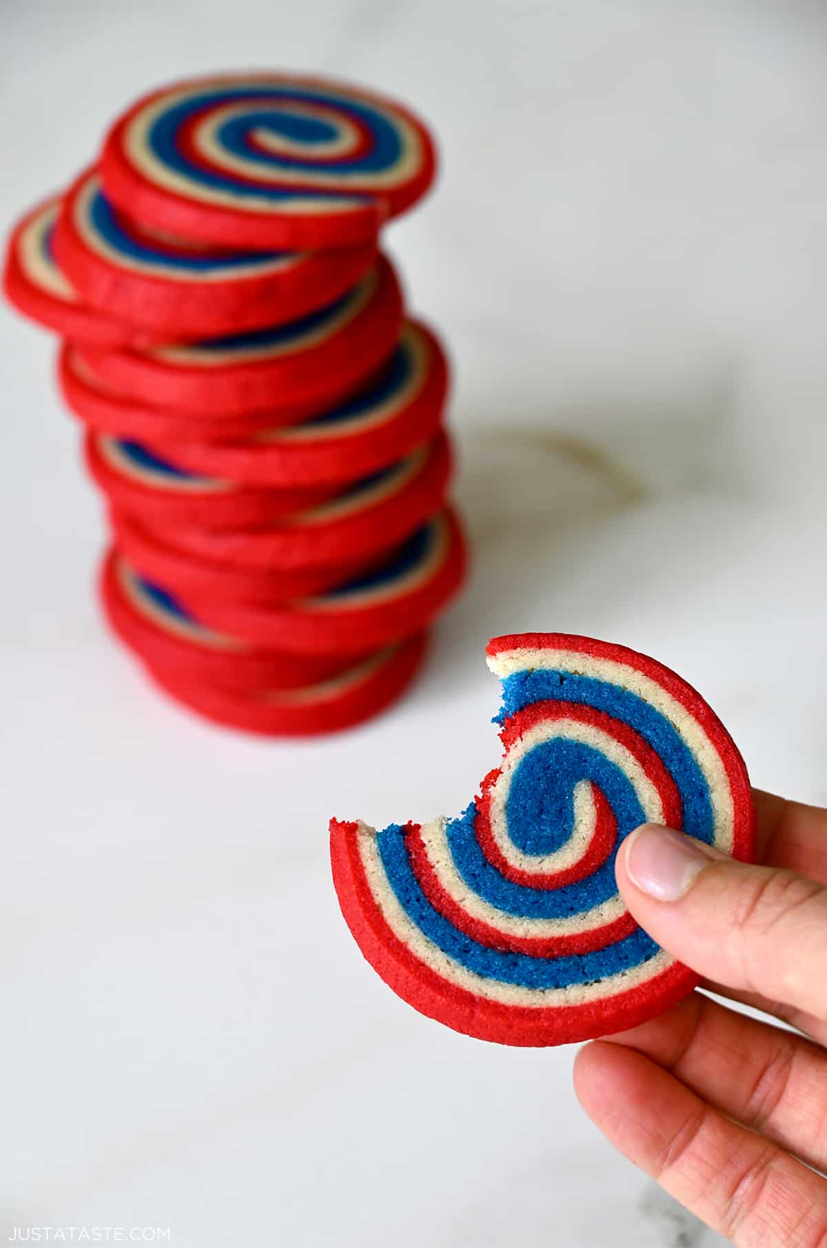 A hand holds a red, white and blue pinwheel cookie with a bite taken out of it in front of a tall stack of pinwheel cookies.