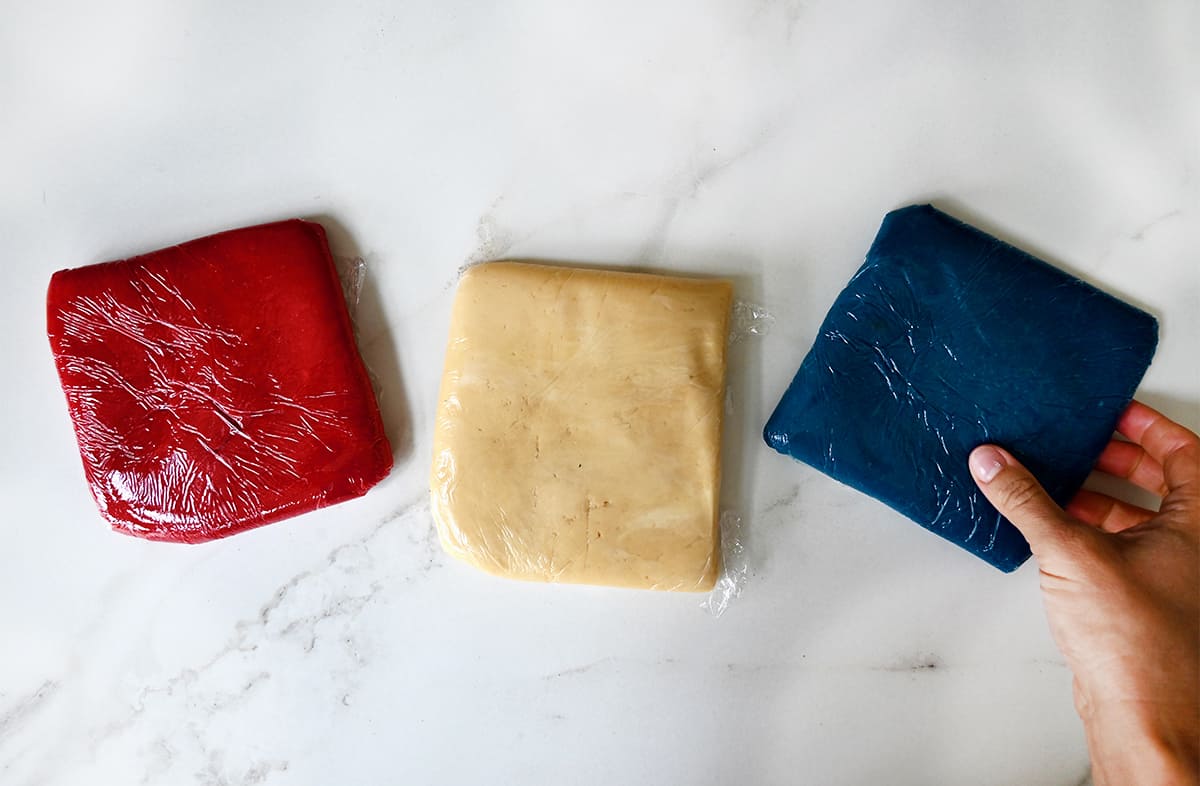 A top-down view of red, white and blue cookie dough wrapped securely in plastic wrap with a hand holding the blue-colored dough.