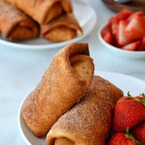 Two Strawberry Cheesecake Chimichangas on a white plate with fresh strawberries