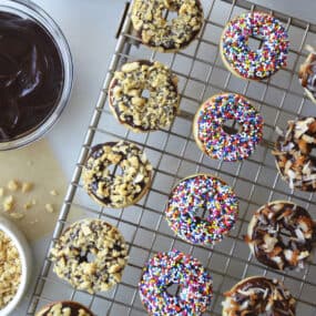Baked Mini Buttermilk Doughnuts atop a wire cooling rack next to bowls containing Nutella Glaze and sprinkles