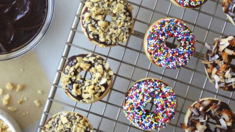 Baked Mini Buttermilk Doughnuts atop a wire cooling rack next to bowls containing Nutella Glaze and sprinkles