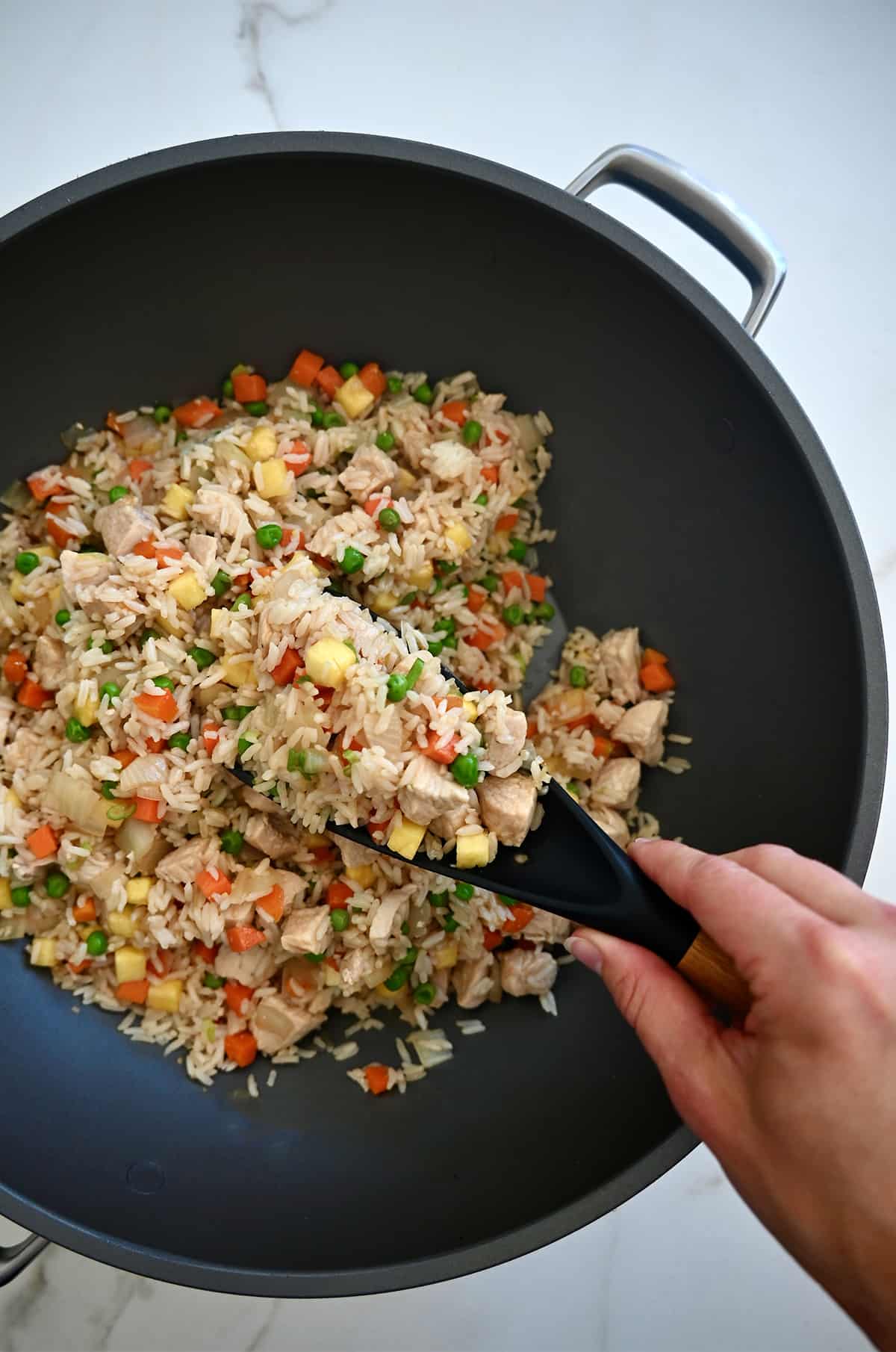 A hand holding a spoon with chicken fried rice over a wok with more fried rice.