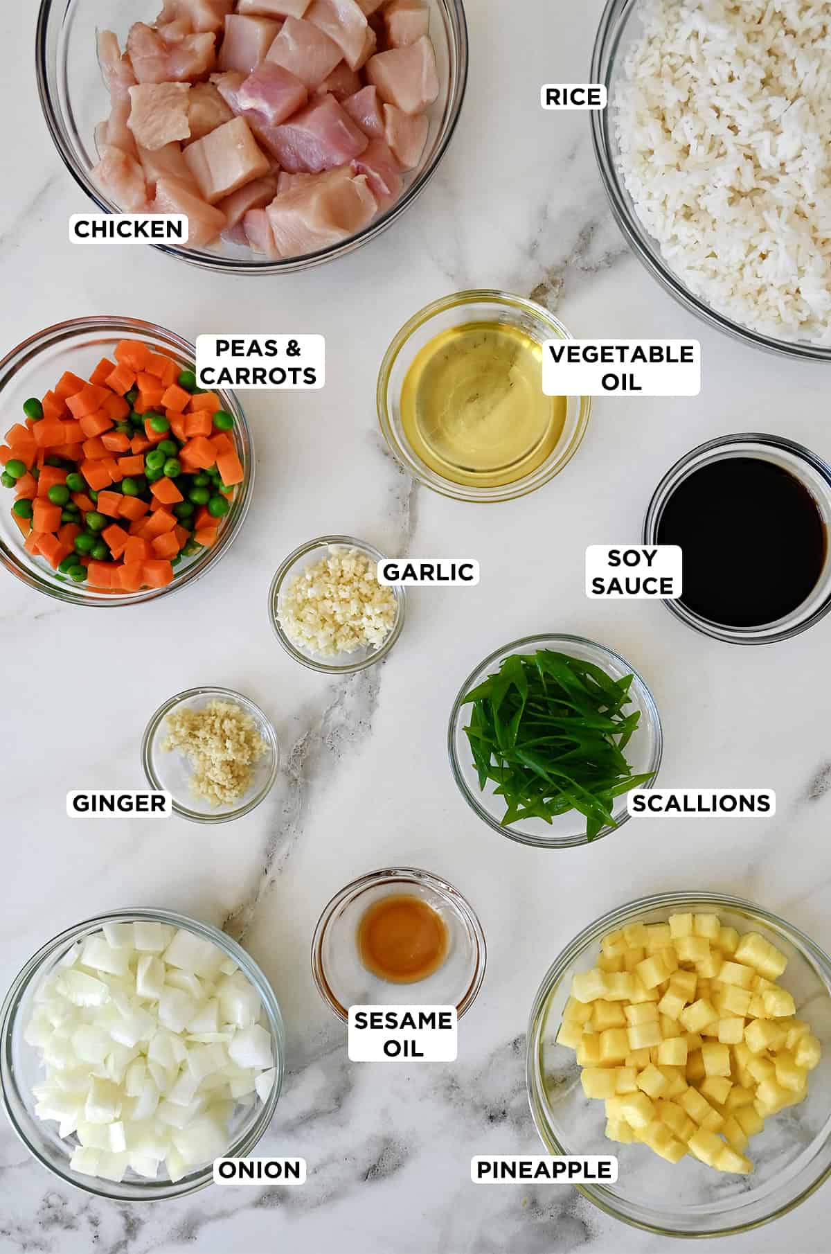Ingredients for pineapple chicken fried rice in various sizes of glass bowls, including diced chicken breasts, day-old rice, vegetable oil, soy sauce, garlic, peas and carrots, scallions, ginger, sesame oil, diced pineapple and onions.