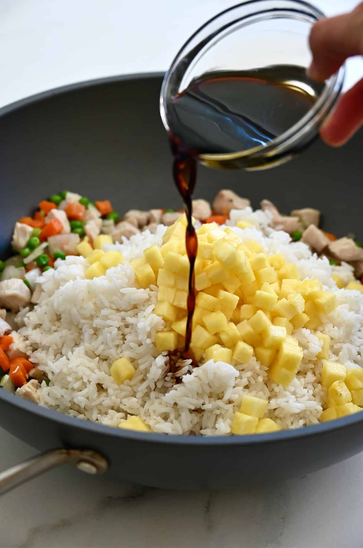 Soy sauce being poured from a small bowl over diced pineapple and rice in a wok.