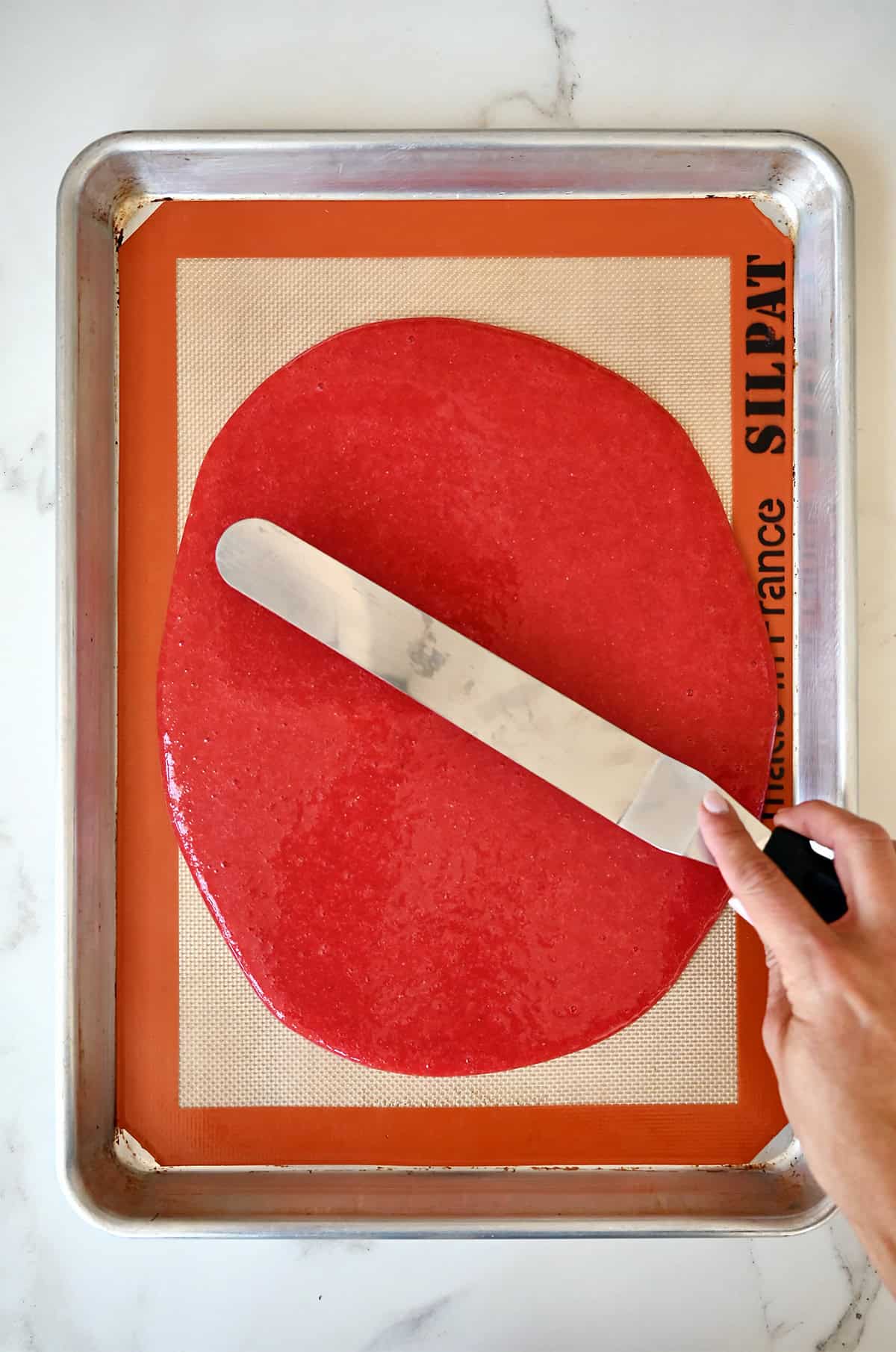 A hand holding an offset spatula spreads strawberry purée atop a baking sheet lined with a silicone baking mat.