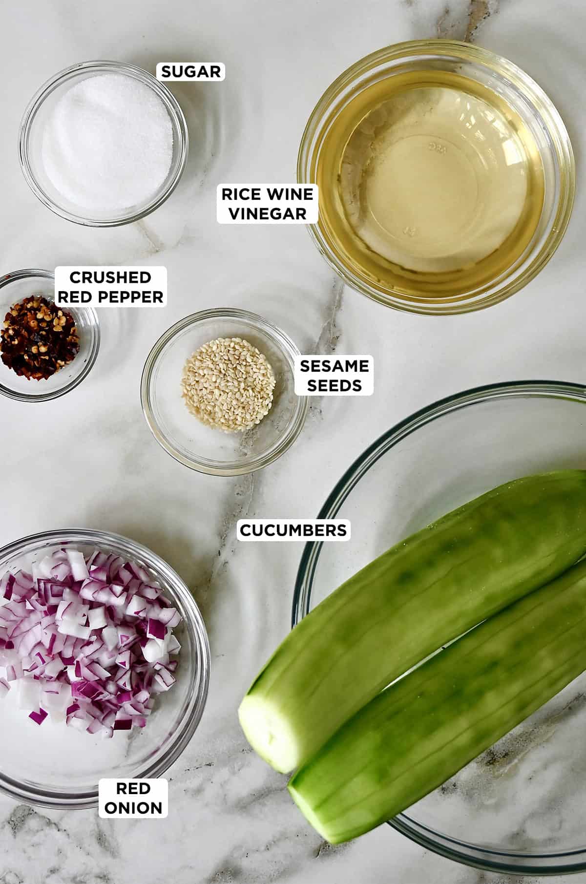Ingredients to make a simple cucumber salad, including two cucumbers, diced red onion, sesame seeds, crushed red pepper flakes, sugar and rice wine vinegar.