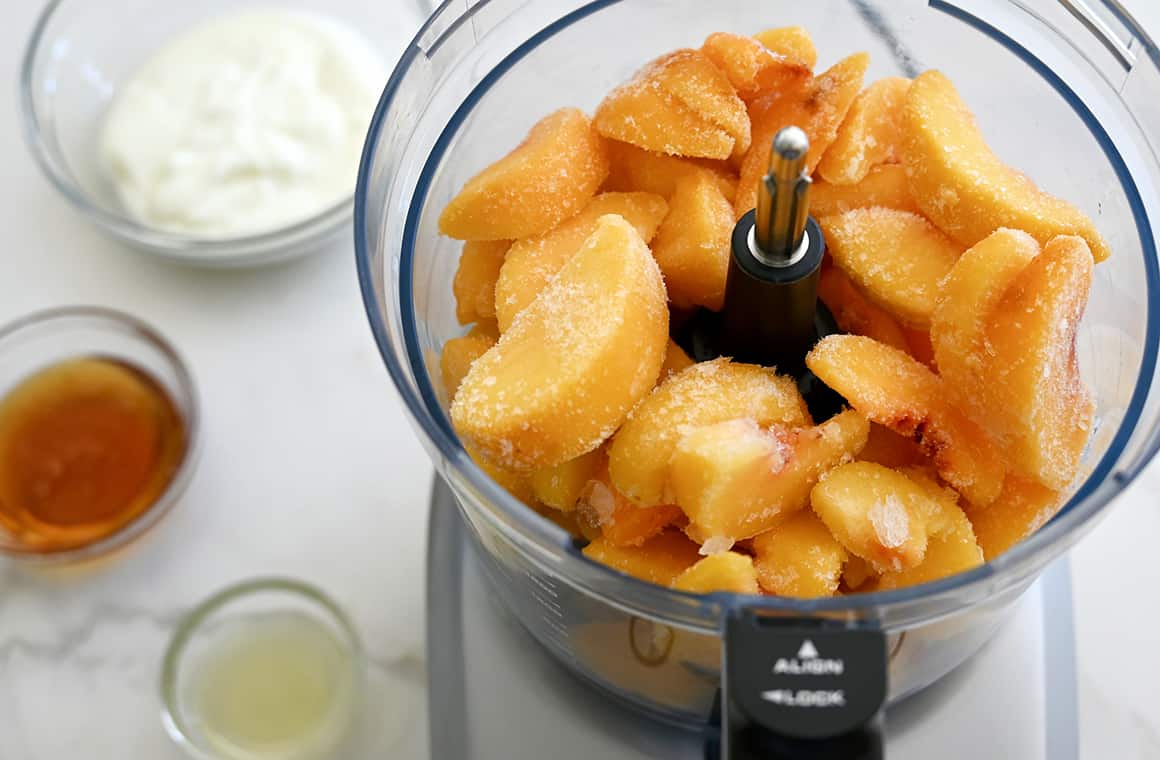 Frozen peaches in a food processor bowl that's next to small bowls containing yogurt, honey and lemon juice