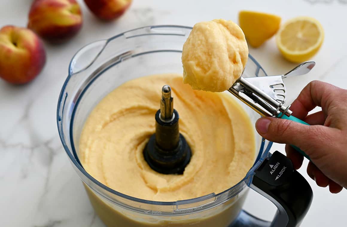 A hand holds an ice cream scoop over the bowl of a food process that contains creamy frozen yogurt