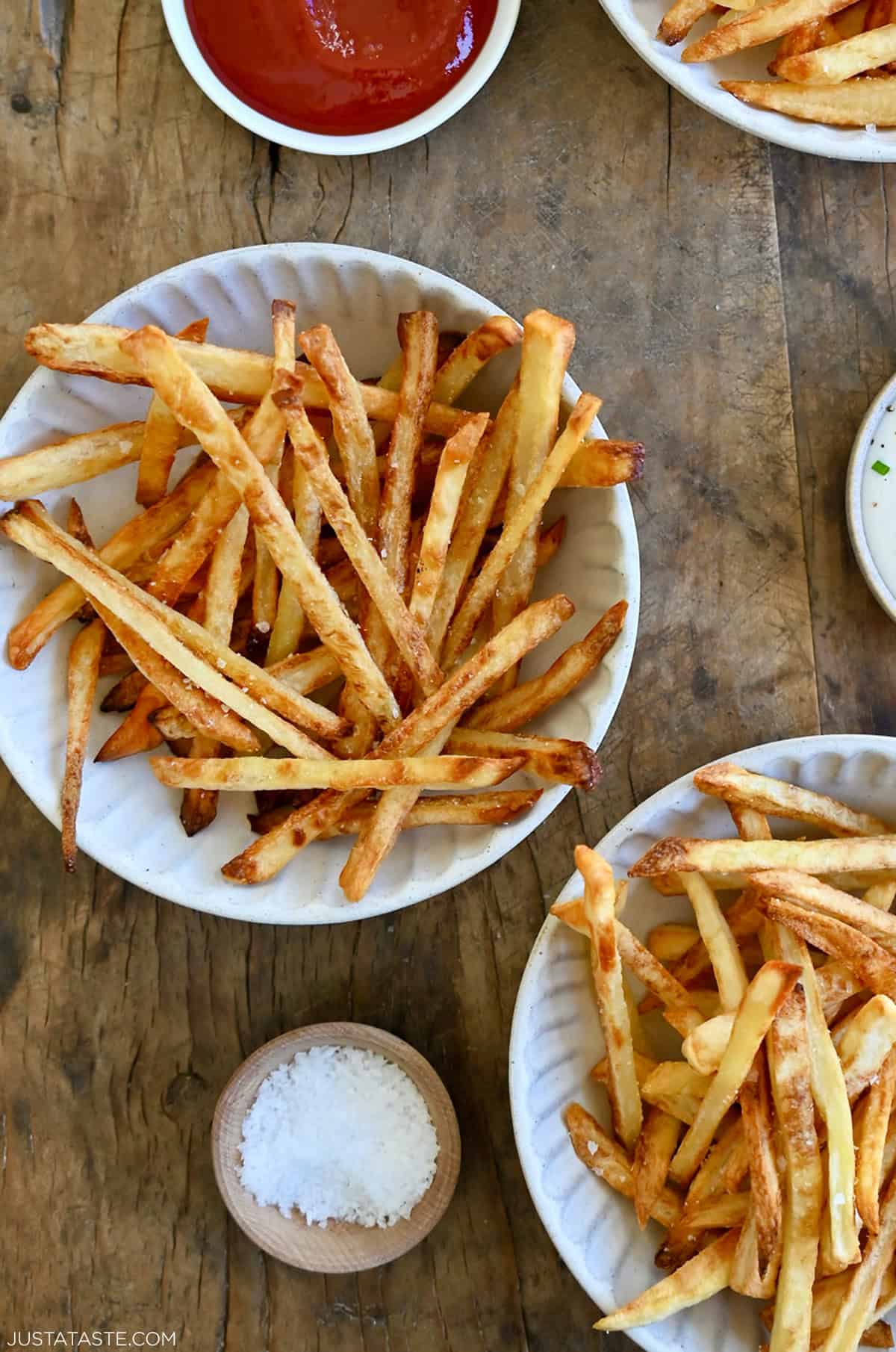 Crispy French fries in two bowls next to a bowl with ketchup, a bowl with sea salt and a bowl with ranch dressing.