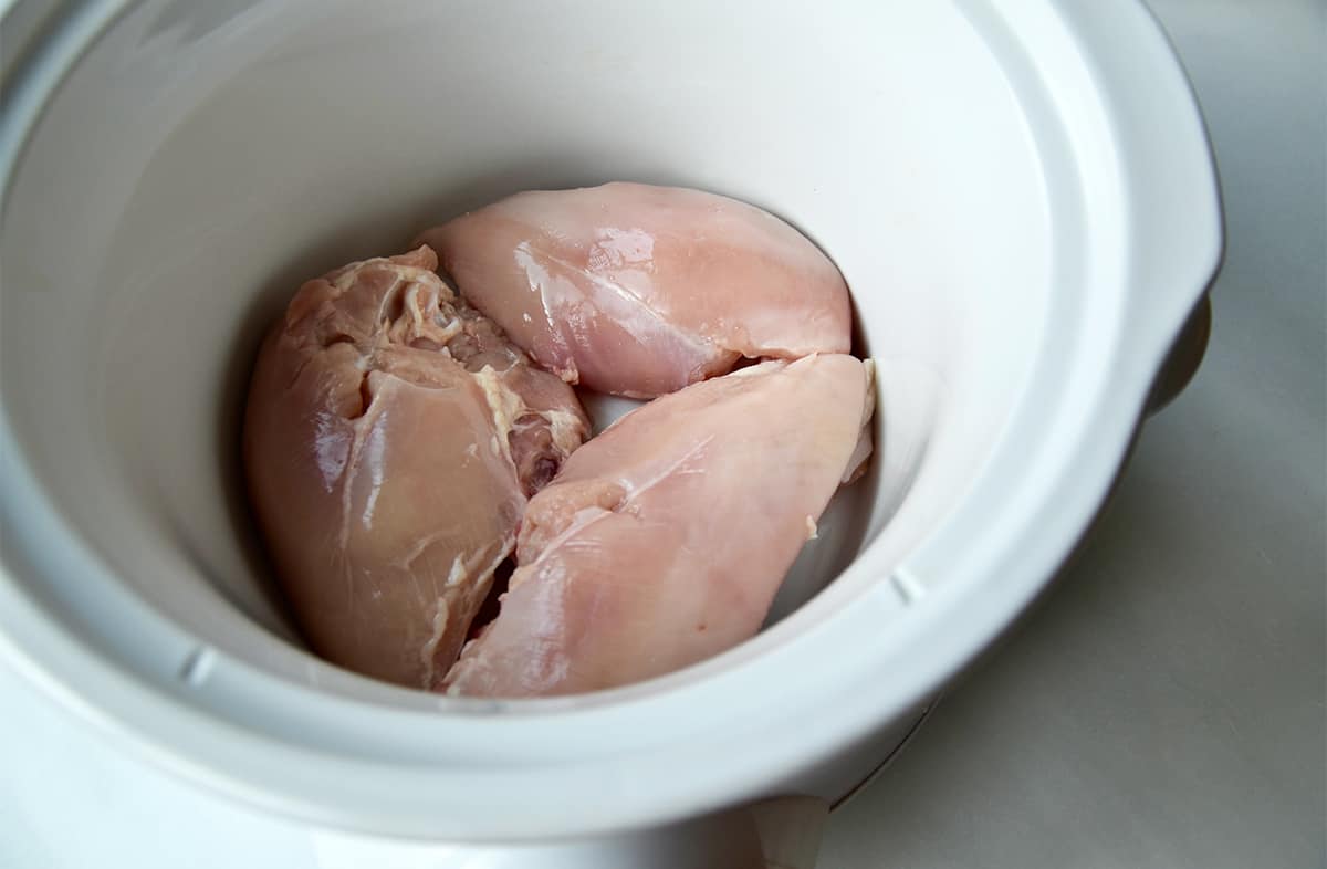 Three raw chicken breasts in a slow cooker.