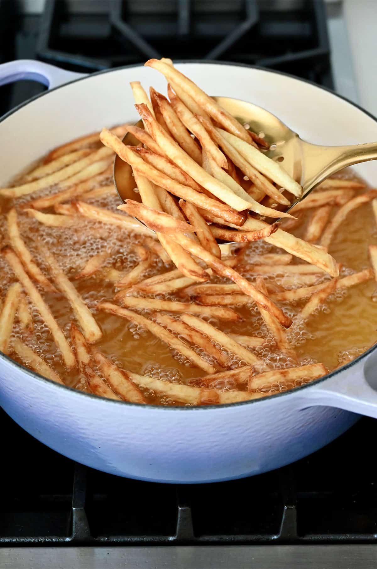 Double-fried French fries on a slotted spoon over a Dutch oven with hot oil and fries cooking.