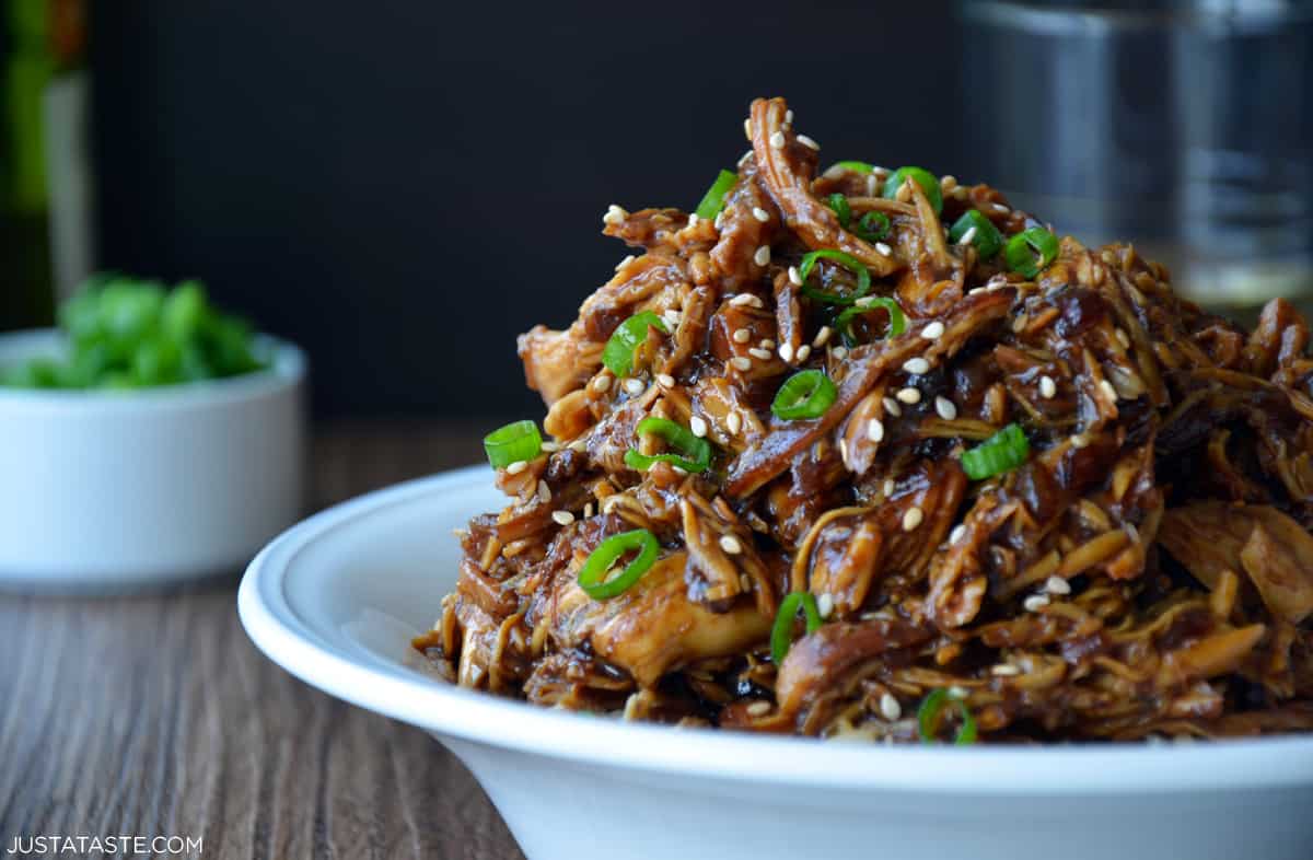 Shredded honey garlic chicken garnished with sesame seeds and sliced scallions over brown rice in a white bowl.