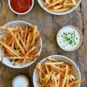 French fries three ways: deep fried, air fried and baked. Three smalls bowls containing ketchup, ranch dressing and sea salt are nearby.