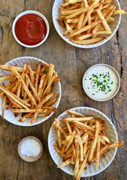 French fries three ways: deep fried, air fried and baked. Three smalls bowls containing ketchup, ranch dressing and sea salt are nearby.