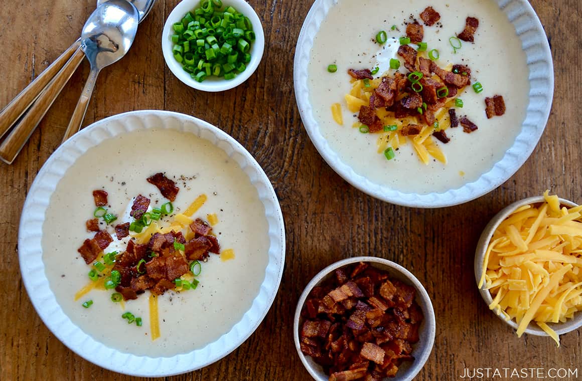 Top-down view of two bowls containing Loaded Baked Potato Soup topped with crispy bacon, sour cream, cheddar cheese and chopped scallions next to three small bowls containing shredded cheddar cheese, chopped bacon and scallions