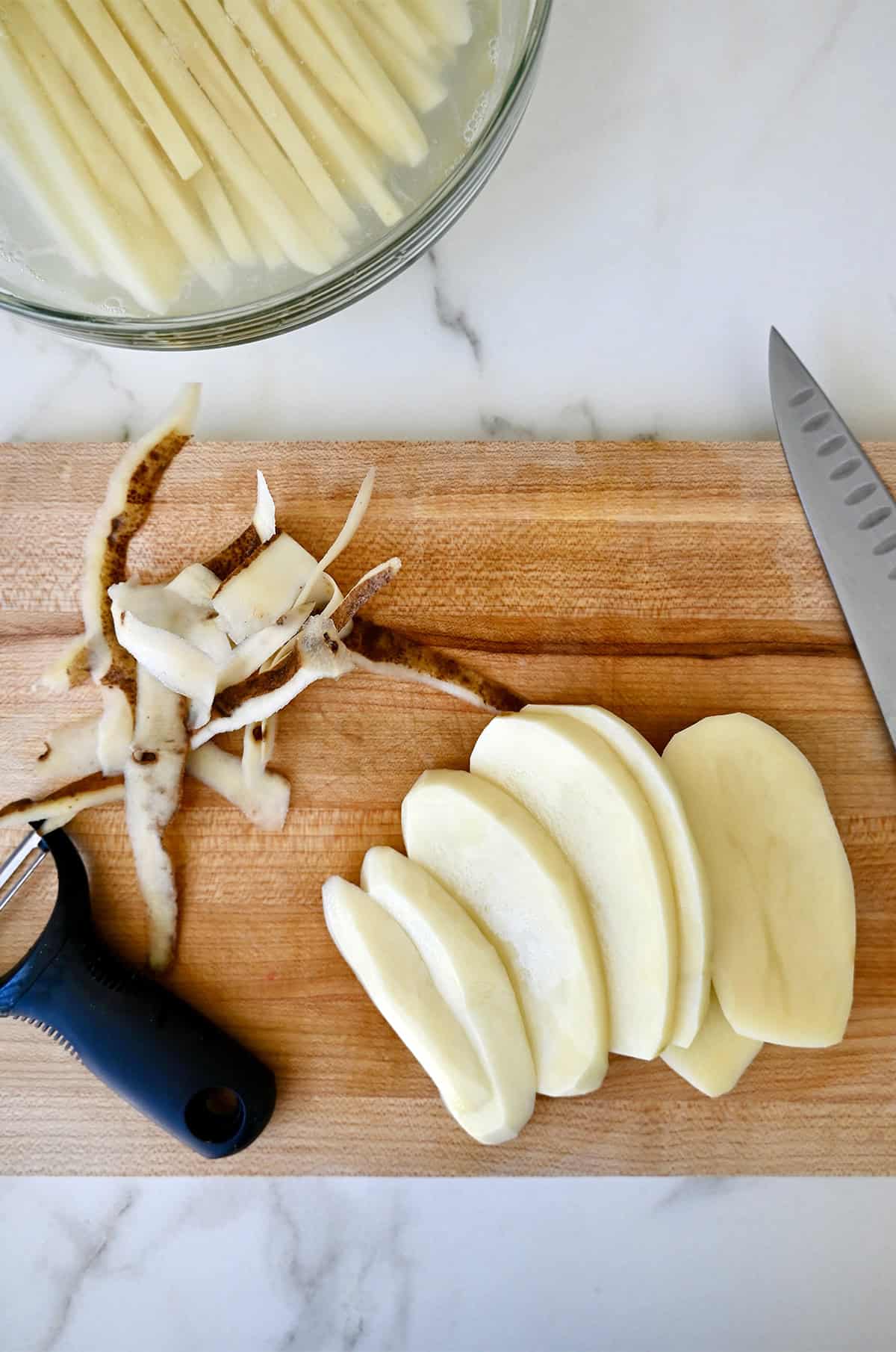 A peeler and a knife on a cutting board with potato skins and a Russet potato cut into slices.