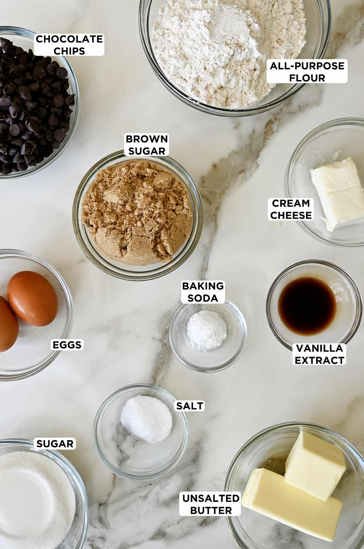 Glass bowls containing ingredients to make soft secret ingredient chocolate chip cookies, including flour, cream cheese, brown sugar, baking soda, vanilla extract, butter, salt, eggs and chocolate chips.