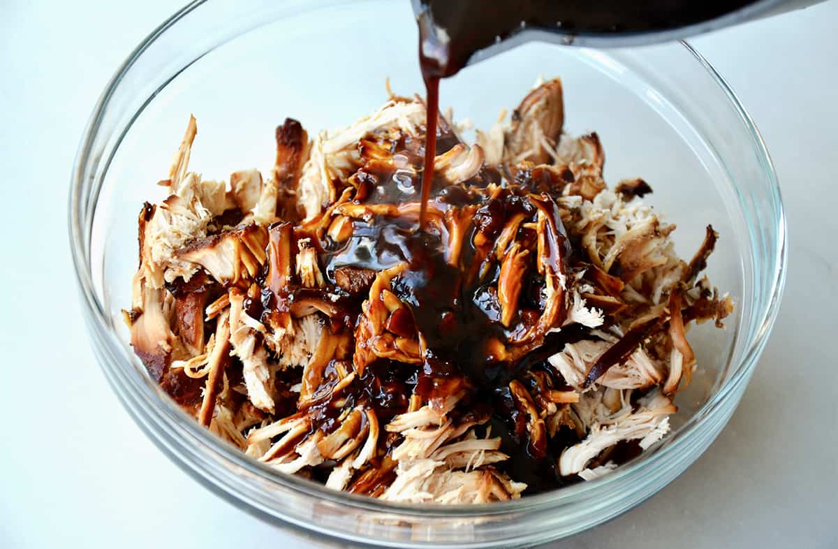 Honey garlic marinade being poured over pulled chicken in a glass bowl.