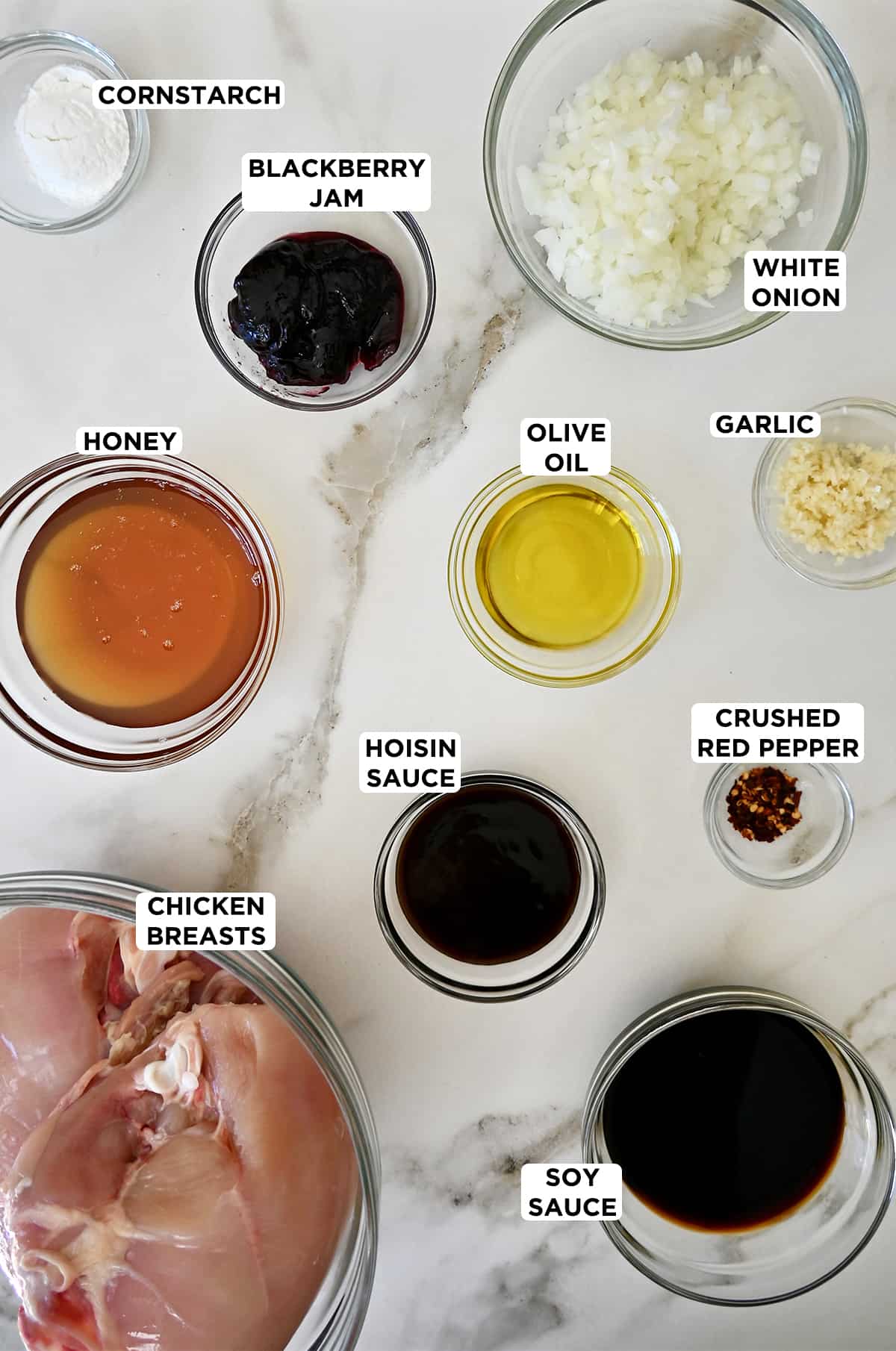 Various sizes of glass bowls containing the ingredients needed to make honey garlic chicken, including cornstarch, blackberry jam, diced white onion, olive oil, minced garlic, crushed red pepper flakes, hoisin sauce, honey, chicken breasts and soy sauce.
