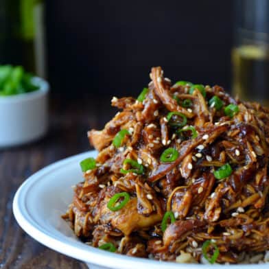 Shredded honey garlic chicken over brown rice in a bowl and garnished with sliced scallions and sesame seeds.