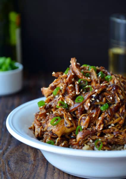 Shredded honey garlic chicken over brown rice in a bowl and garnished with sliced scallions and sesame seeds.